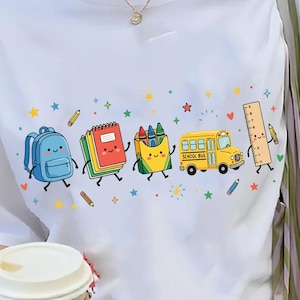 May include: A white short-sleeve t-shirt featuring a colorful graphic print of anthropomorphic school supplies including a blue backpack, a red notebook, a yellow crayon box, a yellow school bus, and a ruler, all surrounded by small stars.