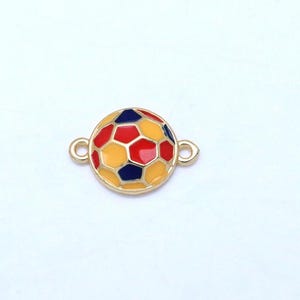 May include: A gold-colored charm in the shape of a soccer ball. The ball features a pattern of red, yellow, and blue pentagons and hexagons. The charm has two small loops on either side for attaching to a bracelet or necklace.