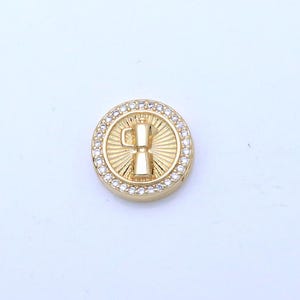 May include: A gold-coloured round charm with a coffee maker design at the centre. The charm is surrounded by a row of small, clear crystals. The background is plain white.