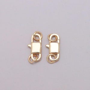 May include: Two gold-toned metal lobster claw clasps, each featuring a rectangular central body with a spring-loaded trigger mechanism and a small circular jump ring attached to both the top and bottom ends of the fastener for jewelry making.