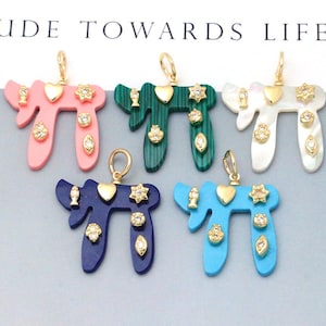 May include: Five Chai pendants in pink, green, white, dark blue, and light blue. Each pendant features a gold-tone metal bail and is decorated with small gold-tone charms including a heart, a star, and clear crystals set in gold-tone metal.