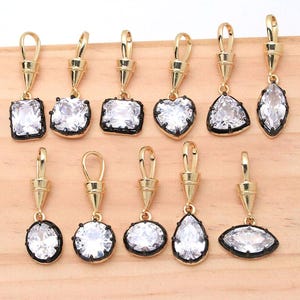 May include: A collection of eleven gold-toned metal pendants, each featuring a clear faceted gemstone set in a black frame. The pendants have a conical bail at the top and come in various shapes including rectangular, heart, round, and pear.