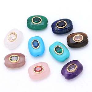 May include: A collection of nine oval-shaped gemstone beads, each featuring a central faceted stone set in a gold-toned bezel. The beads include various colors such as green, blue, brown, purple, pink, and white, with smooth, polished surfaces.