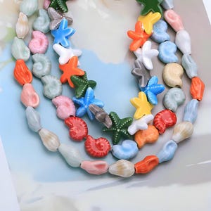 Könnte beinhalten: Eine mehrsträngige Halskette mit bunten Muschel- und Seesternperlen. Die Perlen sind in verschiedenen Farben gehalten, darunter Orange, Rosa, Blau, Grün und Gelb. Die Halskette ist wahrscheinlich ein Accessoire mit Sommer- oder Strand-Thema.