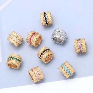 May include: A collection of ten cylindrical beads featuring a textured leaf-like pattern. Most beads are gold-toned with a central vertical row of inlaid stones in various colors, including blue, red, green, black, pink, purple, and turquoise.