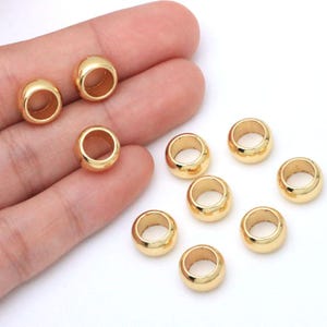 May include: A collection of small, smooth, round gold-colored metal beads with a polished finish. Each bead features a central circular hole, designed for threading. The beads have a reflective surface and a simple, uniform, spherical shape.