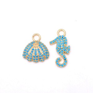 May include: Two gold-toned metal charms featuring a seashell and a seahorse shape. Both charms are encrusted with small, bright turquoise blue stones and include a circular jump ring at the top for attachment to jewelry or other accessories.