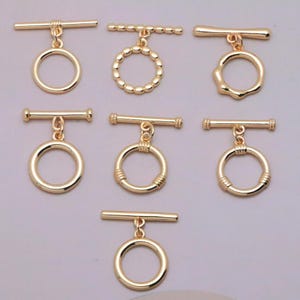 May include: A collection of seven gold-toned metal toggle clasps for jewelry making. Each piece consists of a horizontal bar and a circular ring connected by a small jump ring. The designs vary in texture, including smooth, beaded, and ribbed finishes.
