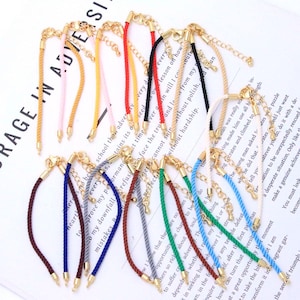 May include: A collection of twelve bracelets featuring twisted rope cords in various colors including red, yellow, pink, black, blue, green, brown, and gray. Each bracelet is finished with gold-toned metal end caps and a lobster claw clasp.