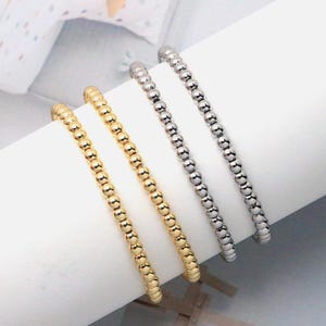 May include: Four beaded bracelets. Two are gold-colored, and two are silver-colored. The bracelets are made of small, round beads strung together. They are displayed on a white, curved surface.