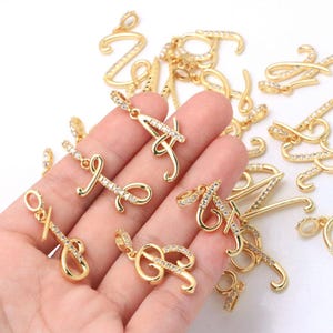 Pode incluir: Uma coleção de pingentes de letras em metal dourado com designs em letra cursiva. Cada pingente é adornado com pequenas pedras de zircônia cúbica claras e brilhantes incrustadas ao longo da superfície das letras. Cada peça inclui uma pequena argola circular.
