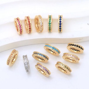 May include: A collection of circular jewellery beads featuring gold-toned metal frames embedded with small, colourful cubic zirconia stones. The beads are arranged in various hues including pink, red, white, green, blue, orange, purple, and black.