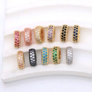 May include: A collection of eleven cylindrical beads featuring gold-tone metal settings encrusted with small, multi-coloured cubic zirconia stones. The beads are arranged in two rows, showcasing various colours including pink, white, purple, black, blue, green, and silver.