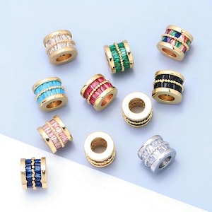 Könnte beinhalten: Eine Sammlung zylindrischer Perlen mit goldfarbenen Metallbändern und zentralen Reihen aus Baguette-geschliffenen Kristallen in verschiedenen Farben, darunter klar, grün, mehrfarbig, türkis, rosa, schwarz, blau und silberfarben, mit Perlenverzierungen.