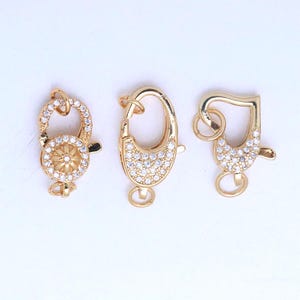 May include: Three gold-colored jewelry clasps with different designs. One clasp is round with a floral center and clear stones. The second is oval-shaped and covered in clear stones. The third is heart-shaped with clear stones.