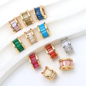 Könnte beinhalten: Eine Sammlung von elf zylindrischen Perlen mit Baguette-geschliffenen Kristallen in verschiedenen Farben wie Blau, Grün, Rot, Rosa und Transparent, eingefasst in goldfarbene Metallrahmen. Jede Perle hat ein Mittelloch zum Auffädeln.