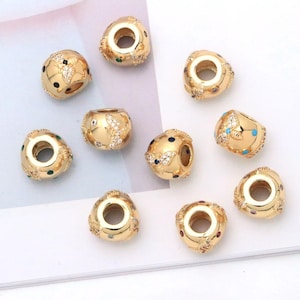May include: A collection of nine gold-toned metal beads, each featuring a central hole and a decorative design of small clear crystals and tiny colored enamel dots. The beads have a smooth, polished finish and a rounded, slightly tapered shape.