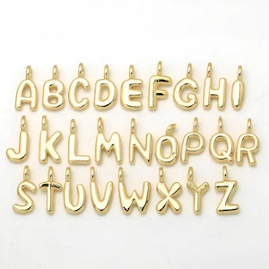 May include: A collection of twenty-six individual gold-colored metal letter charms, arranged in three rows from A to Z. Each letter features a smooth, rounded, puffed surface and includes a small circular loop at the top for attachment purposes.