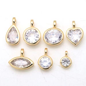 May include: A collection of seven gold-toned metal pendants, each featuring a clear, faceted gemstone set in a bezel. The shapes include a teardrop, two circles, an oval, a heart, and a marquise, all equipped with a small loop for attachment.