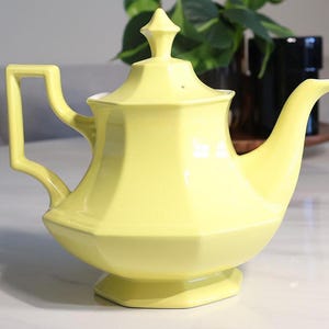 May include: A pale yellow ceramic teapot with a faceted, hexagonal body, a matching lid with a pointed finial, a curved handle, and a tapered spout. The teapot features a wide base and a smooth, glossy finish, presenting a clean, geometric design.