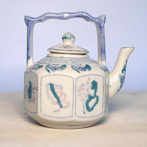 May include: A hexagonal ceramic teapot with a cream-colored glaze, featuring a blue-tinted arched handle and a matching lid. The body is decorated with hand-painted motifs in blue, green, and pink, including floral patterns and stylized insects.