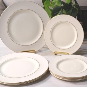 May include: A set of off-white ceramic plates featuring a thin gold rim accent. The collection includes two larger dinner plates and three smaller salad or dessert plates, all arranged with a smooth, circular design and a clean, polished finish.