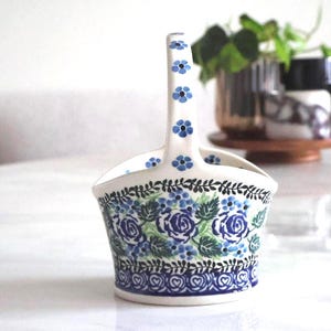 May include: A white ceramic basket with a handle, adorned with blue floral patterns and green leaves. The design includes blue roses and a heart-shaped border. The handle features small blue flower accents. This decorative basket is ideal for home decor.