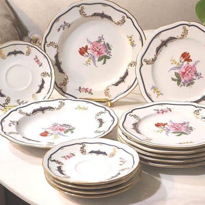 Pode incluir: Um conjunto de louça de porcelana branca com bordas recortadas, acabamento dourado e detalhes em azul escuro. Cada peça é decorada com um motivo floral central em rosa, vermelho e verde, complementado por delicados padrões de ouro.