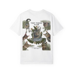 May include: A white t-shirt featuring a graphic print of multiple tabby cats nestled among green tropical leaves and small flowers. The text reads TINY TIGERS CLUB and Sometimes You Forget... We're Tigers. The cats are depicted in various poses.