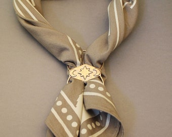 May include: A taupe and white patterned scarf is held together by a gold-colored metal clasp. The scarf features geometric designs and is folded to create a stylish accessory. The clasp has a star and crescent moon design.