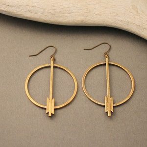 May include: A pair of gold-colored hoop earrings. Each earring features a circular design with a vertical bar and a geometric arrow-like shape at the bottom. The earrings have a hook closure.