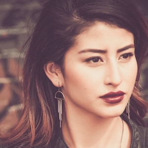 May include: A woman with dark hair and burgundy lipstick wears a black leather jacket and silver earrings. The earrings have a half-circle design with dangling chains. The background is a blurred brick wall.