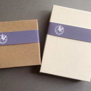 May include: Two rectangular boxes, one brown and one off-white, are displayed on a gray surface. Each box is wrapped with a lavender band featuring a white logo of a bird within a circle. The boxes are likely for packaging.