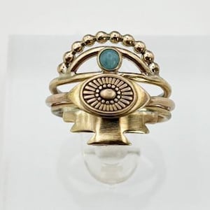 May include: A gold-toned ring stack featuring three distinct bands. The top band has a beaded arch with a small blue stone. The middle band displays an oval eye-shaped centerpiece with radiating lines. The bottom band has a geometric fan shape.