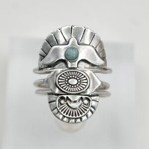 May include: A silver ring featuring three stacked horizontal segments. The top segment displays a bird motif with a central round light blue stone. The middle segment is an oval with radial lines, and the bottom segment is a semi-circle with rays.