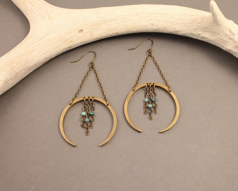 May include: A pair of brass-toned earrings with a crescent moon design. Each earring features a chain that connects to a circular frame, with turquoise beads dangling from the center. The earrings have a hook closure.