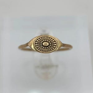 May include: A gold-toned ring featuring an oval-shaped centerpiece with a central raised dot surrounded by a ring of small beads and a radiating sunburst pattern of thin, elongated lines, all set within a smooth, tapered oval frame.