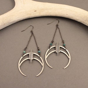 May include: Silver-toned earrings with a crescent moon design. Each earring features a chain, a vertical bar with three lines, and two curved lines. Small turquoise beads are incorporated. The earrings hang from a hook.