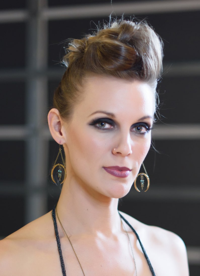 May include: A woman with dark eye makeup and a nose ring is wearing gold-colored crescent moon earrings with turquoise accents. The earrings hang from a chain. Her hair is styled in a bun.