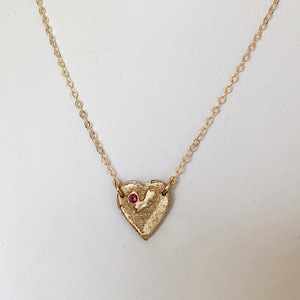 Peut inclure: Un collier en chaîne dorée avec un petit pendentif en forme de cœur texturé orné d'une seule pierre précieuse rouge.