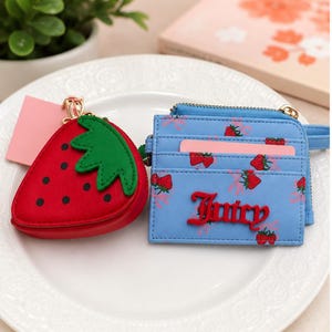 May include: A red strawberry-shaped coin purse with green leaf detailing and black dots. Beside it is a light blue cardholder, patterned with strawberries and the red word "Juicy".