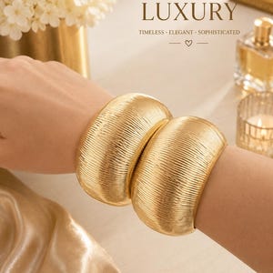 May include: Two wide gold-toned cuff bracelets featuring a textured, ribbed surface. The bracelets have a rounded, convex shape and a polished metallic finish. The text LUXURY, TIMELESS, ELEGANT, SOPHISTICATED is visible in the upper right corner.