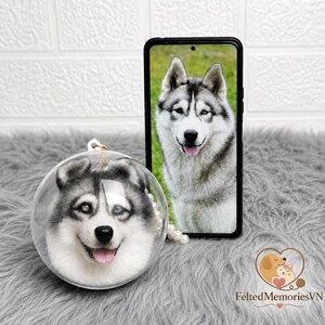 以下が含まれることがあります： 笑顔のフェルトハスキー犬の顔が付いた透明な丸いオーナメント。スマートフォンには同様のハスキーの画像が表示されています。オーナメントは灰色の表面にあり、真珠のネックレスと「FeltedMemoriesVN」のロゴが付いています。