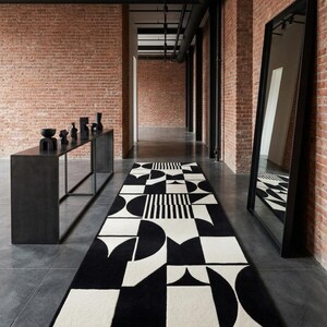 May include: A long rectangular runner rug featuring a bold black and white geometric pattern of circles, squares, and stripes. The rug is positioned on a dark floor in a room with exposed brick walls, a black console table, and a tall floor mirror.