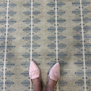 Könnte beinhalten: Ein gewebter Teppich mit einem sich wiederholenden Muster aus vertikalen Streifen und blauen blattartigen Motiven auf hellbeigem Grund. Der Teppich hat eine strukturierte Oberfläche und ist mit einem Paar hellrosa Slipper am unteren Rand zu sehen.