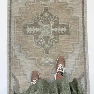 Könnte beinhalten: Ein rechteckiger Teppich mit einem zentralen geometrischen Medaillonmuster in gedämpften Beige-, Taupe- und sanften Grautönen. Der Teppich hat eine strukturierte, gewebte Optik mit einem symmetrischen Design und einem einfachen Rand.