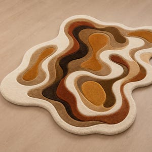 Könnte beinhalten: Ein unregelmäßig geformter Teppich mit einem geschichteten, topografischen Design und organischen, fließenden Konturen. Der Teppich zeigt eine Farbpalette aus Creme, Beige, Goldgelb, Braun und Dunkelschokolade, was einen multidimensionalen Effekt erzeugt.