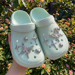 May include: A pair of light mint green slip-on clogs featuring silver-toned charms. The charms include detailed butterflies, four-pointed stars, smooth spheres, and a heart shape attached to the ventilation holes on the top of the footwear.