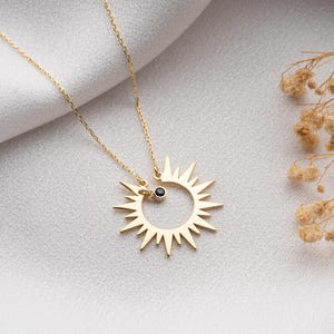 May include: A gold-colored necklace featuring a sunburst pendant with a small black gemstone. The pendant is attached to a delicate chain. The necklace is displayed on a white, textured surface, with dried flowers in the background.