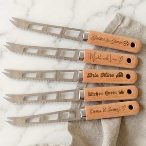 Könnte beinhalten: Fünf Käsemesser mit silberfarbenen Metallklingen, rechteckigen Aussparungen und gezackten Kanten. Jedes Messer hat einen hellen Holzgriff mit den Gravuren: Gather & Graze, Made with Love, Brie Mine, Kitchen Queen und Emma & James.